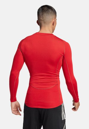 Uomo con capelli corti di spalle, indossa una maglia sportiva rossa a maniche lunghe aderente e pantaloncini neri con strisce bianche.