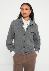 Kobieta w szarej swetrze zapinanym na guziki, nosząca białą koszulę w paski i brązowe spodnie, stojąca z rękami w kieszeniach na tle białej ściany.