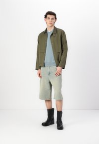 Olive green jacket with snap closures, light blue sweatshirt underneath, light denim shorts, and black ankle boots; casual outfit displayed.
