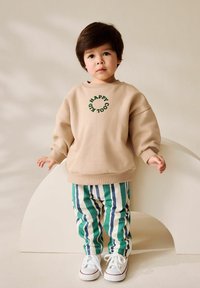 Kleinkind mit dunklen Haaren, das einen beigen Pullover mit dem Text "HAPPY COOL KID", gestreifte grüne und blaue Hose sowie weiße Sneaker trägt, steht drinnen.