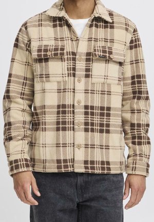Beige und braunes kariertes Fleece-Hemd zum Knöpfen mit zwei geknöpften Brusttaschen, über einem weißen Unterhemd und dunklen Jeans getragen.