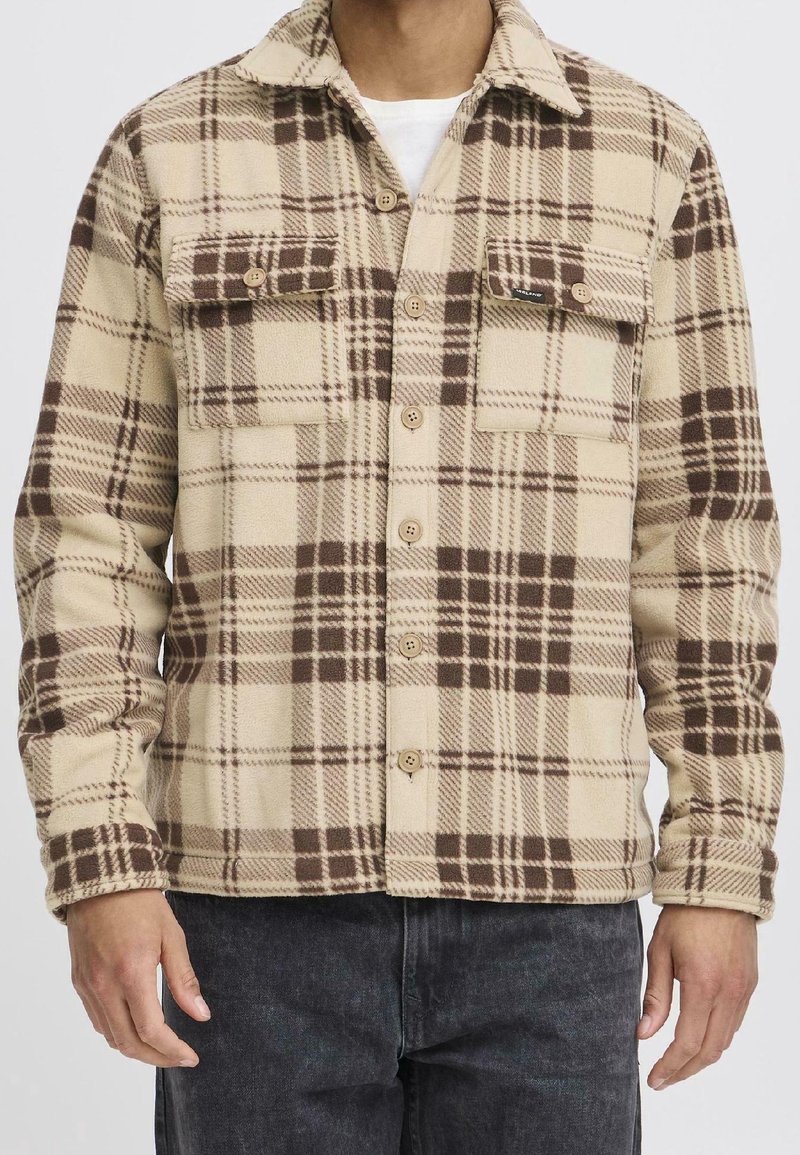 Chemise polaire à carreaux beige et marron à boutons avec deux poches poitrine boutonnées, portée sur un maillot blanc et un jean foncé.
