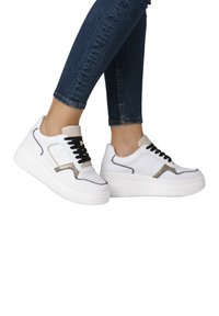 Weiße Sneakers mit schwarzen Schnürsenkeln, kontrastierenden Nähten in Schwarz und Beige sowie einer dicken weißen Sohle. Das Design umfasst Mesh-Akzente und eine gepolsterte Zunge.