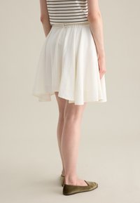 Falda blanca hasta la rodilla con un dobladillo acampanado, hecha de tela texturizada; combinada con zapatos planos de color verde oliva. Parte de una blusa a rayas es visible.