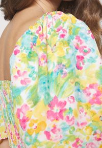 Gros plan sur l'épaule d'une femme portant une robe à manches bouffantes florale colorée avec des motifs roses, jaunes, verts et bleus sur un tissu blanc.