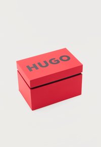 Κόκκινο ορθογώνιο κουτί με μαύρο κείμενο "HUGO" στο καπάκι, τοποθετημένο σε απλό ανοιχτό γκρι φόντο.