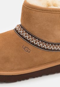 Bota de tobillo de ante marrón con una superficie texturizada, que presenta una banda con diseño trenzado en marrón, crema y marrón claro. Logotipo mostrado en el lateral.
