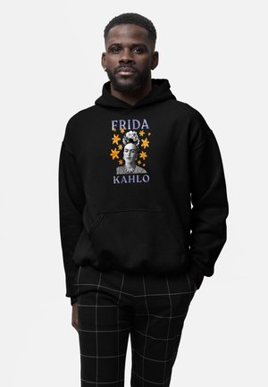 Man draagt een zwarte hoodie met een portret van Frida Kahlo en gele bloemen, gecombineerd met een zwarte broek met witte rasterlijnen.