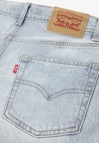 Ανοιχτό μπλε τζιν παντελόνι με ορθογώνια πίσω τσέπη, κίτρινες ραφές και καφέ δερμάτινη ετικέτα με το λογότυπο της μάρκας και κείμενο. Ορατή κόκκινη ετικέτα.