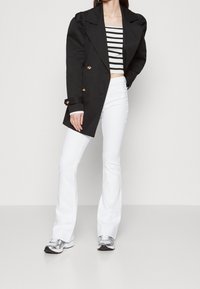 Manteau noir croisé avec des boutons dorés, porté sur un haut cropped à rayures. Assorti à un jean évasé blanc et des chaussures de sport argentées.