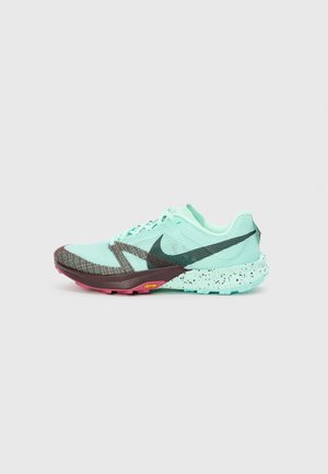 Chaussure de course turquoise clair et marron foncé avec semelle tachetée et logo Vibram, présentée sur un fond gris clair uni.