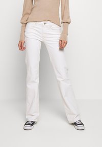 Jeans blancos acampanados de mezclilla de algodón con un bolsillo frontal visible, botón metálico y detalles de costura. Combinados con zapatillas negras de lona.