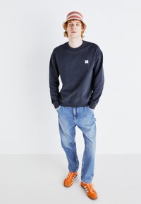 Navy sweatshirt with crew neck, light blue denim jeans, and orange sneakers with stripes. Model wearing a multi-colored striped bucket hat.