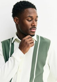 Grün-weiße, gerippte Knopfshirt mit vertikalen Streifen, einem Kragen und strukturiertem Stoff, getragen von einer Person mit nachdenklichem Ausdruck.