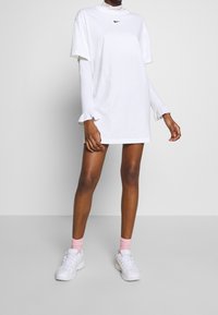 White oversized t-shirt dress with high collar and Nike logo, layered over a fitted long-sleeve shirt with ruffled cuffs. Paired with white sneakers.