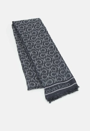 Bufanda gris oscuro con patrón circular repetitivo en gris más claro y texto "Calvin Klein" en el borde con flecos, colocada plana sobre fondo blanco.