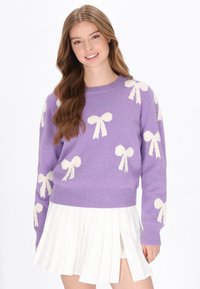 Lila gestrickter Pullover mit weißen Schleifenmustern, Rundhalsausschnitt und gerippten Bündchen. Kombiniert mit einem weißen, plissierten Rock.