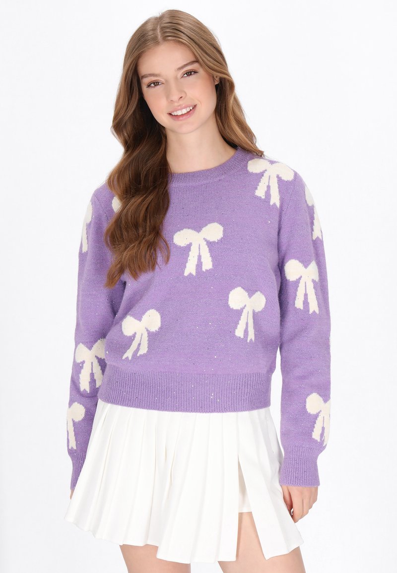 Lila gestrickter Pullover mit weißen Schleifenmustern, Rundhalsausschnitt und gerippten Bündchen. Kombiniert mit einem weißen, plissierten Rock.