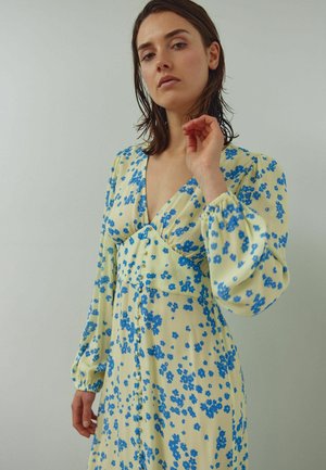 Rochie florală galbenă cu flori albastre, având un decolteu adânc în V, mâneci bufante și nasturi frontali. Material ușor, fluid.