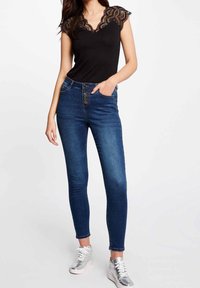 Musta pitsikoristeinen yläosa V-päällä, jonka parina tummansiniset tiukat farkut korkealla vyötäröllä ja napilla kiinnitys, yhdistettynä hopeisiin lenkkareihin.