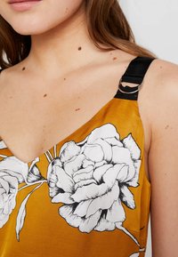 Camisola floreale in giallo senape, caratterizzata da grandi stampe di peonie nere e bianche. Spalline nere con anelli decorativi. Tessuto dalla trama liscia.