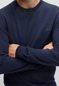 Pull en maille bleu marine avec col rond, manches longues et logo discret "BOSS" sur la poitrine. Texture lisse et coupe ajustée.