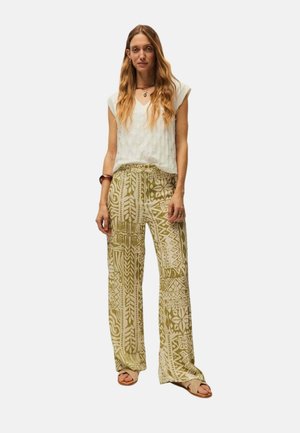 Mujer con cabello largo y ondulado, que lleva una blusa blanca sin mangas, pantalones beige de pierna ancha con estampado, sandalias y joyas sencillas, de pie frente a un fondo liso.