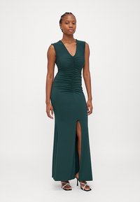 Form-fitting green maxi dress with a V-neck, ruched detailing, and a front slit. Smooth fabric with a sleek finish. High-heeled sandals worn.