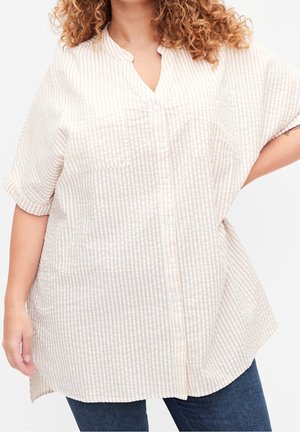 Persoon met krullend haar die een beige en witte verticaal gestreepte kortemouwen blouse met knopen en blauwe spijkerbroek draagt, met hand op de heup.