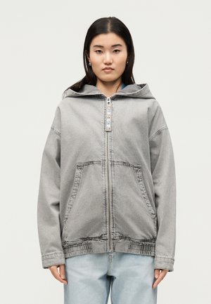 Jonge vrouw met een lichtgrijze capuchonjas met ritssluiting en voorzakken en lichtblauwe spijkerbroek, staand tegen een effen achtergrond.