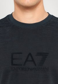 T-shirt azul escuro feito de um tecido suave, com decote redondo e um logotipo preto em relevo que diz "EA7 Emporio Armani" no peito.