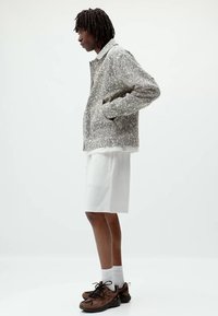 Veste en polaire grise et blanche texturée, vue de côté. Associée à un short blanc, des baskets brunes et des chaussettes blanches. Design minimaliste, sans motifs.