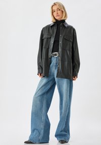 Schwarze Lederhemdjacke mit zwei vorderen Taschen, silbernen Druckknöpfen, kombiniert mit hellblauen weiten Jeans und schwarzen Schuhen.