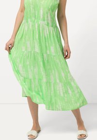 Zelené šaty s vrstveným designem a lehkým abstraktním vzorem, vyrobené z lehkého materiálu. Mají bezrukávový střih a jemné detailní nařasení.