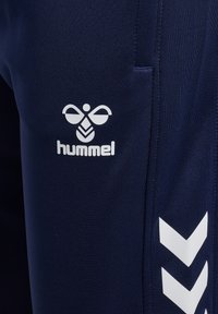 Close-up van marineblauwe sportbroek met wit Hummel-logo en twee witte chevrons langs het zijnaad.