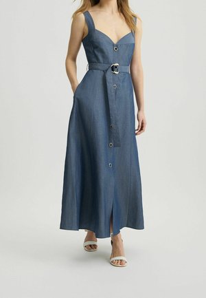 Mujer con vestido largo azul sin mangas abotonado y cinturón en la cintura, con sandalias blancas abiertas, de pie sobre un fondo claro.