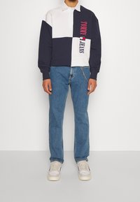 Långärmad polo skjorta i vitt, marinblått och ljusblått med ett färgblockeringsdesign. Har "TOMMY JEANS" text i rosa. Avslappnade denimjeans.