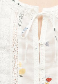 Λαιμός λευκής μπλούζας με δαντελένιο τελείωμα, υφασμάτινες κορδέλες, κουμπιά και τυπωμένα μικρά σχέδια φρούτων και ψαριών.