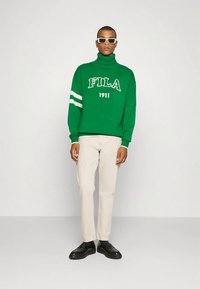 Green knit turtleneck with white "FILA 1911" logo, featuring two white stripes on sleeves. Paired with light beige pants and black shoes.