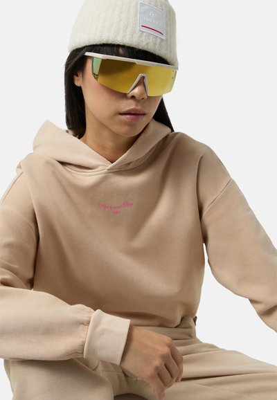 Beżowa bluza z luźnym krojem i różowym haftowanym napisem. Model nosi oversize'owe, odblaskowe żółte okulary przeciwsłoneczne oraz białą dzianą czapkę.