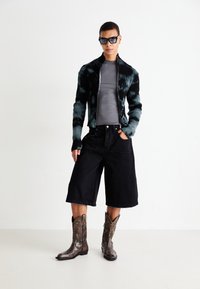 Persoană îmbrăcată cu un hanorac cu fermoar în nuanțe de negru și albastru tie-dye, camasă gri ajustată, pantaloni scurți negri largi, cizme de cowboy maronii cu model și ochelari de soare închiși la culoare.