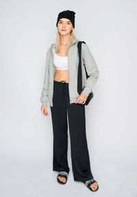 Felpa grigia chiara con zip, top corto bianco, pantaloni joggers neri larghi e sandali neri con un cinturino decorato. Cappellino nero e borsa a spalla inclusi.