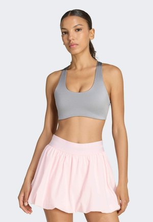 Mujer con el pelo recogido, lleva un sujetador deportivo gris y una falda plisada rosa claro sobre un fondo liso y claro.