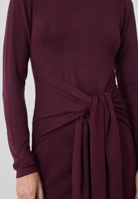 Bordeaux lange mouw jurk met een gedraaide taille detail, zachte stof en een gladde textuur. Knoopontwerp voegt visuele interesse toe.