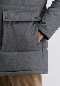 Graue gepolsterte Jacke mit einer großen Fronttasche und Knopfverschluss, strukturiertem Stoff und elastischen Bündchen, kombiniert mit einer braunen Cordhose.