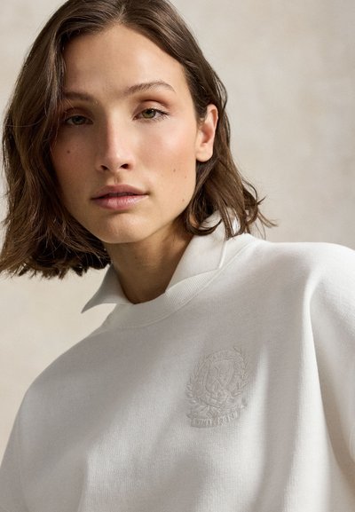 Jeune femme aux cheveux courts et bruns portant un pull blanc avec un blason en relief sur la poitrine, regardant doucement la caméra.
