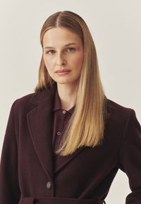 Burgundská vlněná bunda s klopou, jedním knoflíkem a připevněným páskem. Pod ní burgundová košile s límečkem a detaily na knoflících.