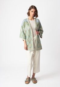 Kimono leggero e decorato in tonalità verdi e pastello, adornato con disegni intricati. Abbinato a pantaloni color crema e sandali.
