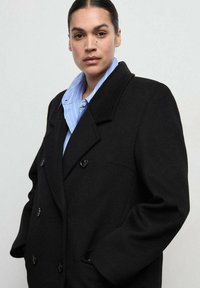 Czarny dwurzędowy płaszcz z dużymi klapami, z dwoma zewnętrznymi kieszeniami i okrągłymi czarnymi guzikami, noszony na niebieskiej koszuli w paski.