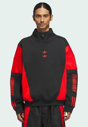 Man with long dark hair in black and red Adidas half-zip sweatshirt with "CHAVARRIA" text on sleeves and matching black and red pants.